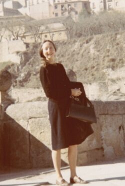 Sepia-toned photo of a woman standing in front of a hillside. Her hair is in a low ponytail and she is wearing a long sleeved black cardigan, black pencil skirt, loafers, and holding a black jacket and black purse. 